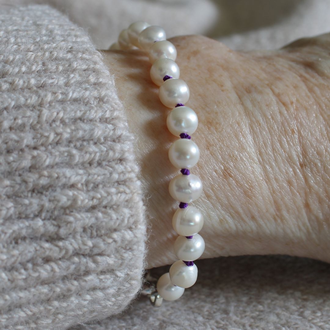 A close-up of a pearl  bracelet hand-knotted with amethyst silk worn on a person's wrist, next to a light gray knit sweater cuff.