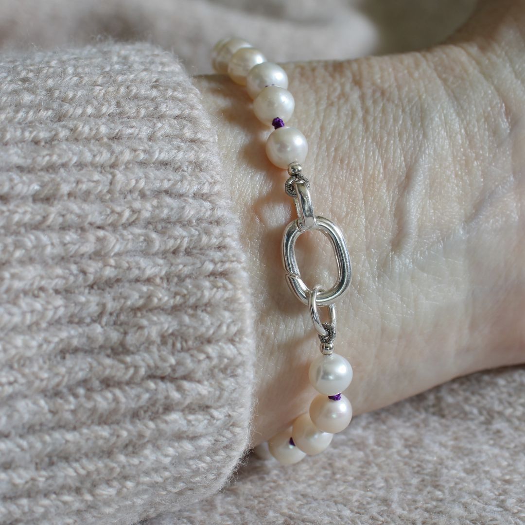A close-up of a pearl beaded bracelet, hand-knotted with amethyst silk, with a silver oval charmer clasp worn on a wrist, resting against a beige knit sweater.