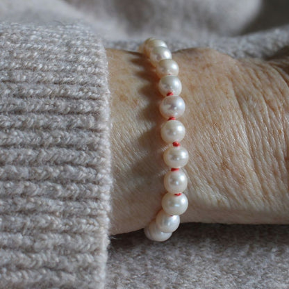 A close-up of a pearl bracelet, hand-knotted on coral silk, worn on a person's wrist over a light gray knit sweater.