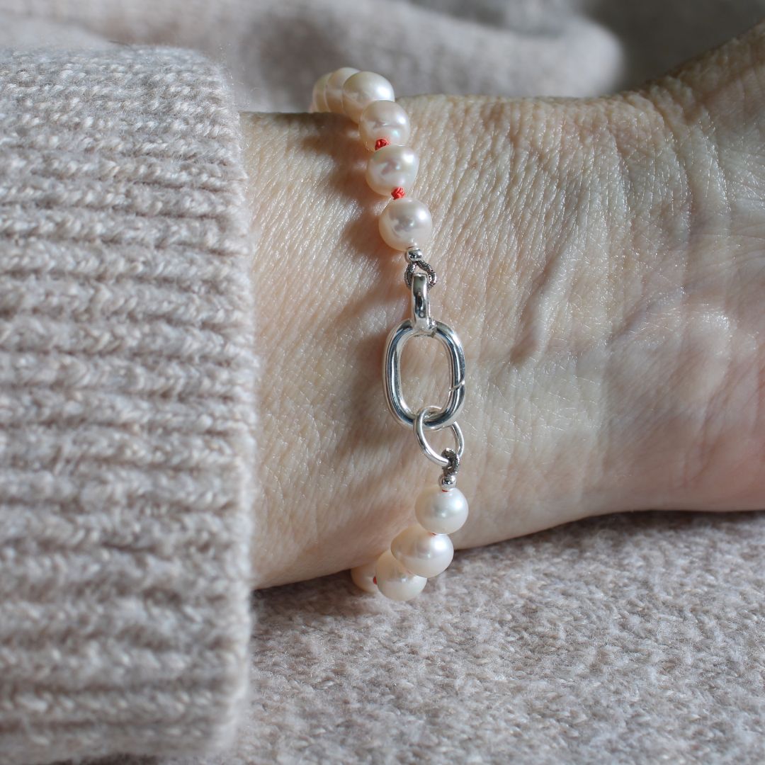 A close-up of a person's wrist wearing a bracelet made of white pearls knotted on coral silk and a silver oval charm clasp.