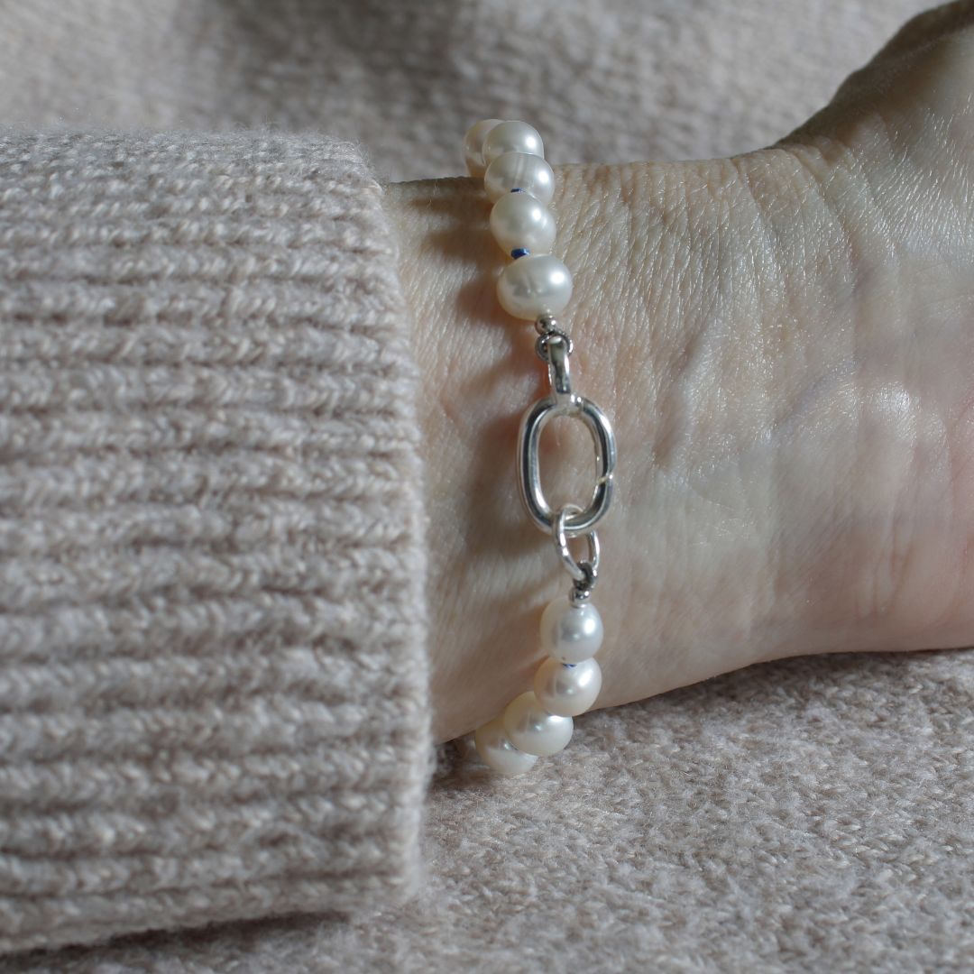 A close-up of a pearl bracelet with a silver oval link clasp worn on a wrist, styled with a beige knit sweater cuff.