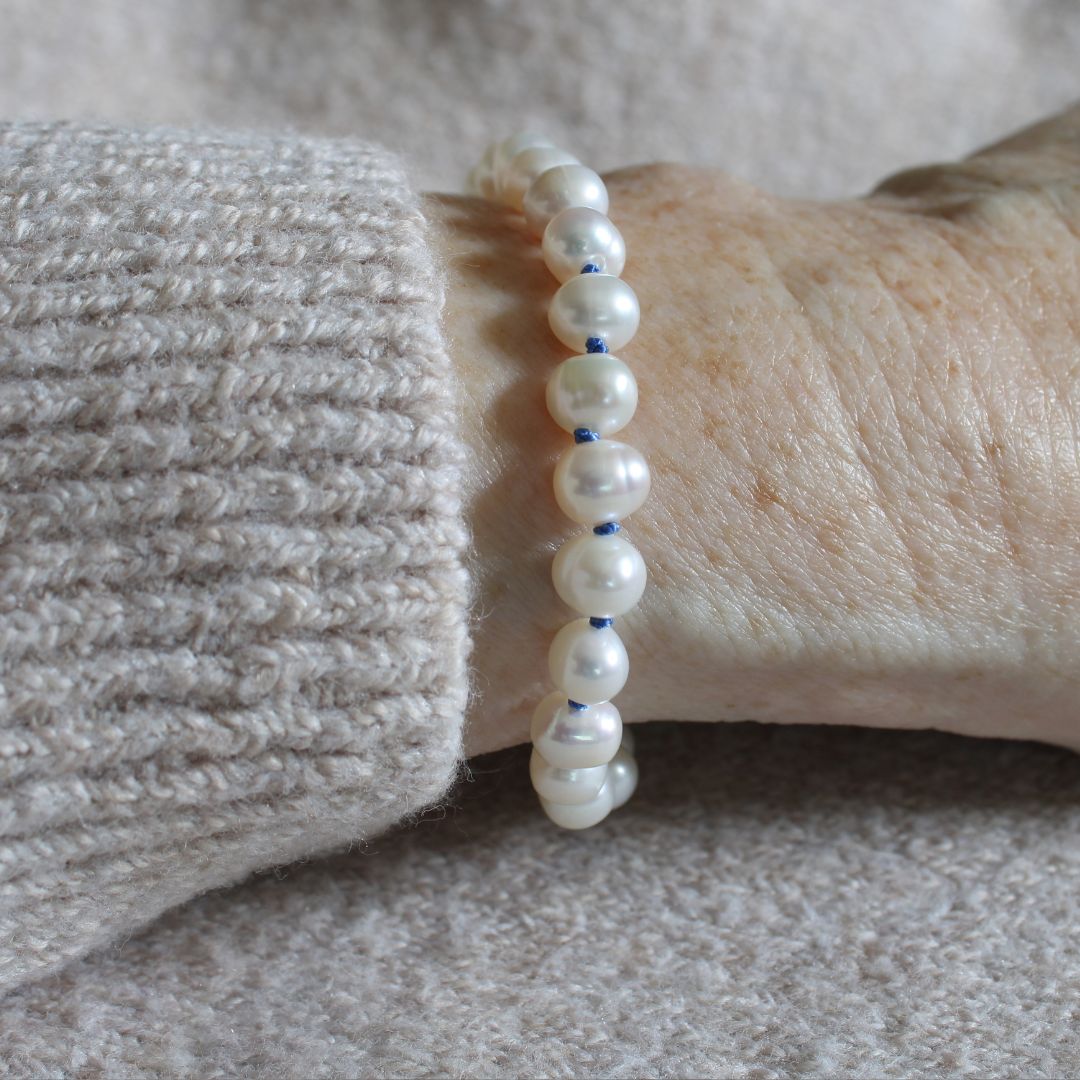 A close-up of a person's wrist wearing a beaded bracelet made of white freshwater pearls knotted with delft blue silk, paired with a beige knit sweater cuff.