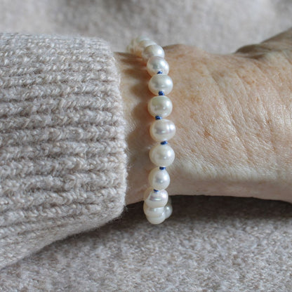 A close-up of a person's wrist wearing a beaded bracelet made of white freshwater pearls knotted with delft blue silk, paired with a beige knit sweater cuff.