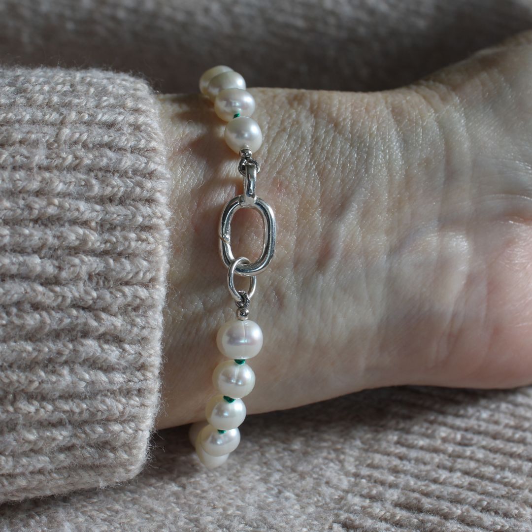 A close-up of a person's wrist wearing a bracelet made of white freshwater pearls and a polished silver clasp, paired with a beige knit sweater.