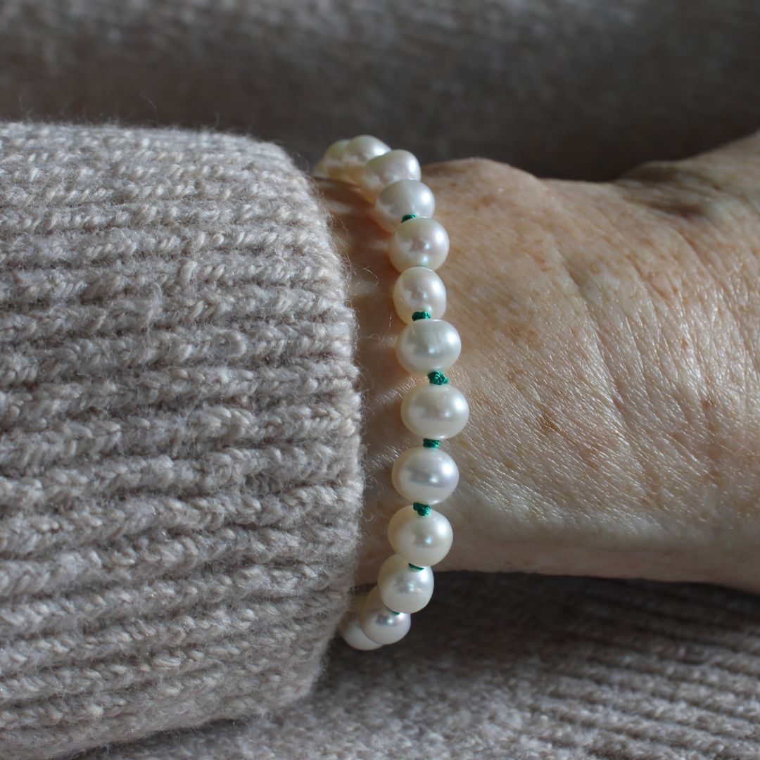 A close-up of a beaded bracelet with white pearls knotted on emerald green silk, worn on a person's wrist over a ribbed beige sweater.