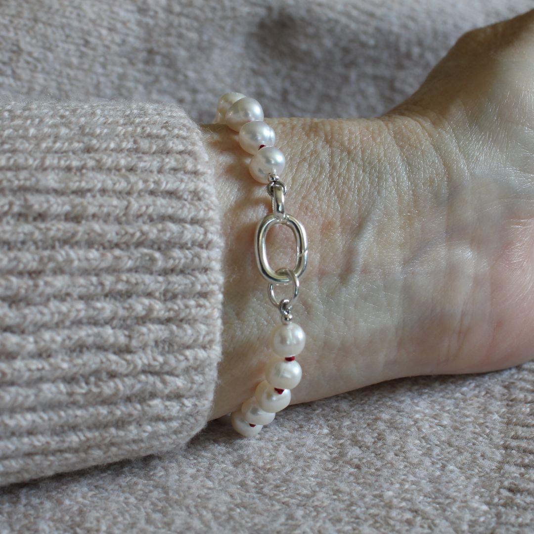 A close-up of a pearl bracelet, knotted with garnet silk, with a silver oval charmer clasp worn on a wrist, next to a ribbed beige knit sleeve.