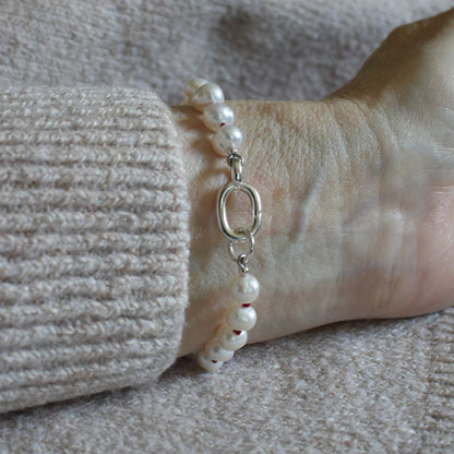 A close-up of a pearl bracelet, knotted with garnet silk, with a silver oval charmer clasp worn on a wrist, next to a ribbed beige knit sleeve.