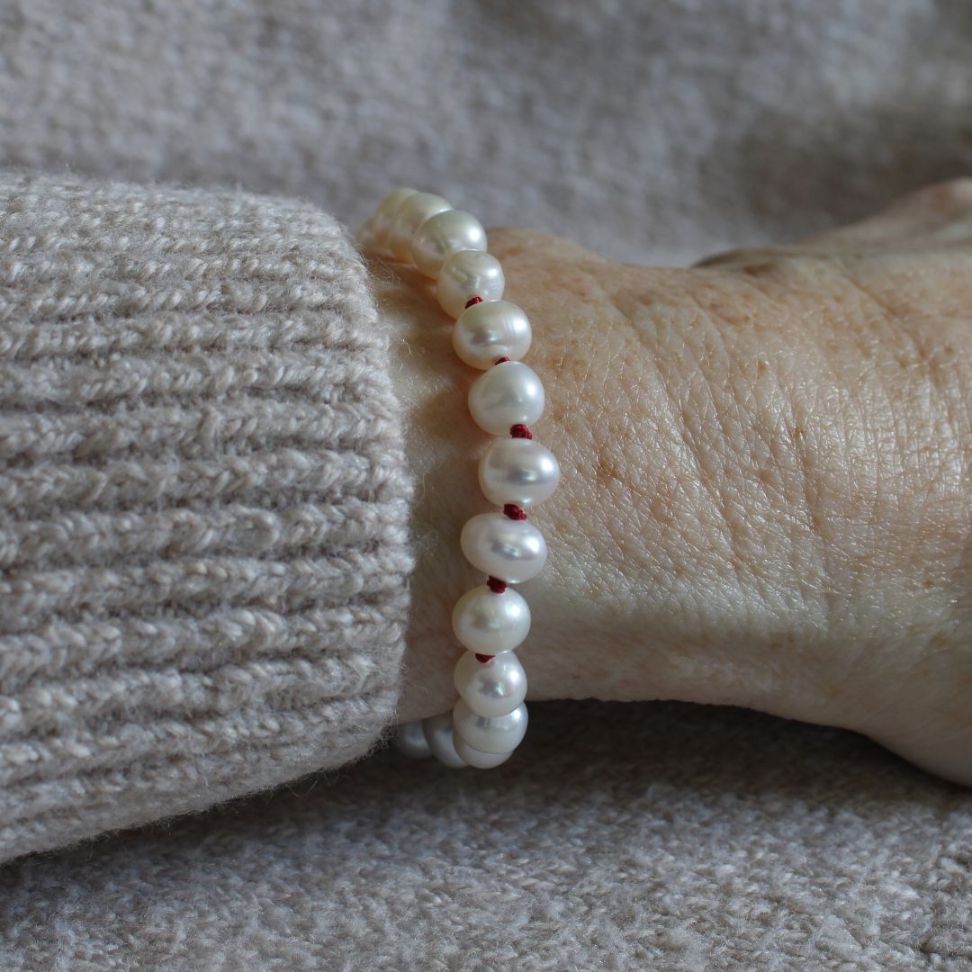 A close-up of a person's wrist pearl bracelet hand-knotted with garnet silk, worn with a beige ribbed knit sweater sleeve.