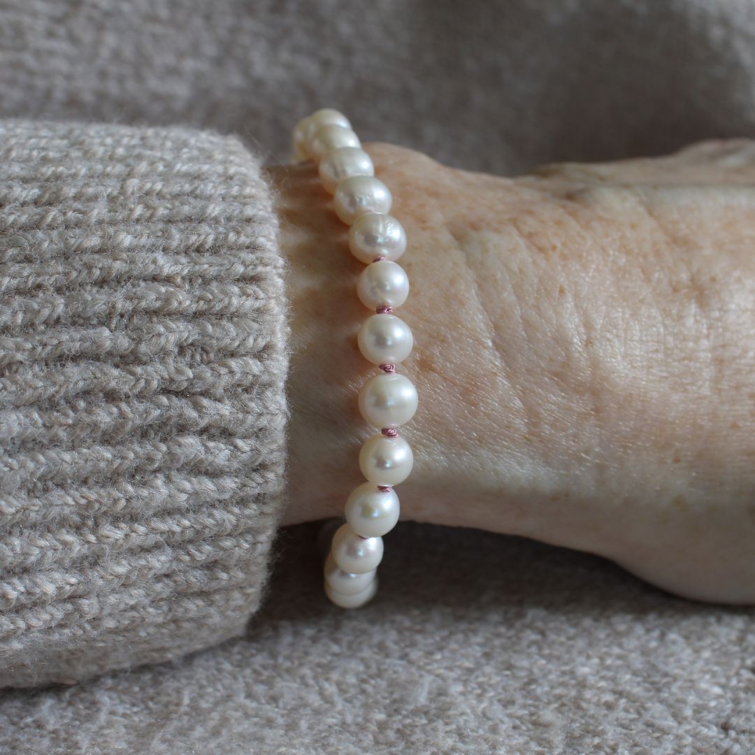 A close-up of a person's wrist wearing a beaded bracelet made of white pearls knotted with old rose silk, paired with a beige knit sweater cuff.