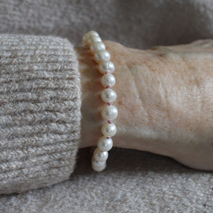 A close-up of a person's wrist wearing a beaded bracelet made of white pearls knotted with old rose silk, paired with a beige knit sweater cuff.