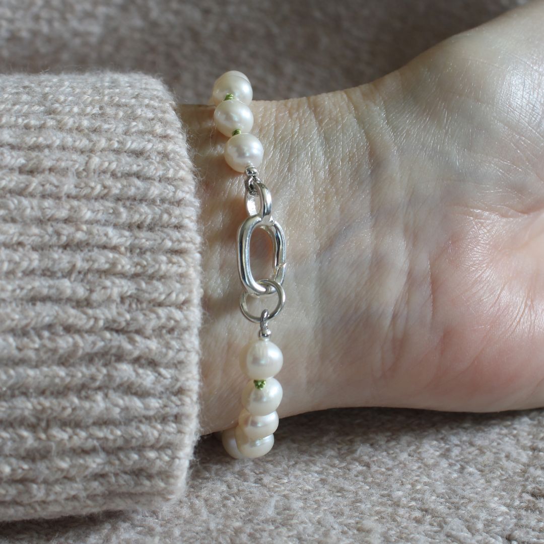 A close-up of a person's wrist wearing a bracelet made of white freshwater pearls and a silver clasp, with a beige knit sweater cuff visible.