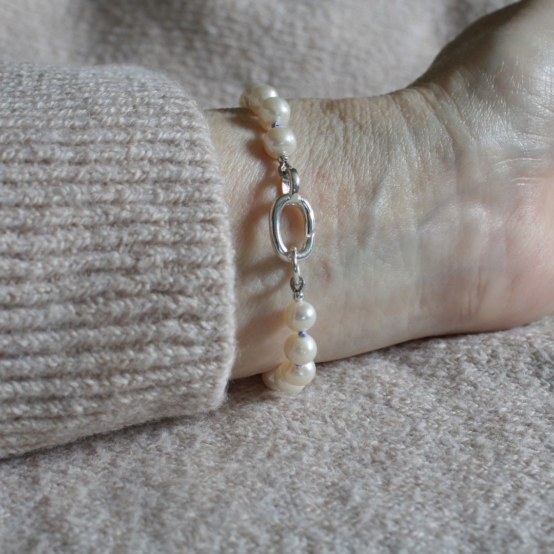 A close-up of a pearl bracelet with periwinkle silk and silver oval charmer clasp, worn on a wrist with a beige ribbed sweater cuff visible.