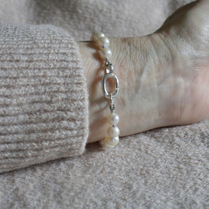 A close-up of a pearl bracelet with periwinkle silk and silver oval charmer clasp, worn on a wrist with a beige ribbed sweater cuff visible.