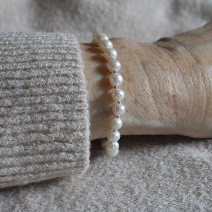 A close-up of a person's wrist wearing a pearl bracelet made white pearls, knotted with periwinkle silk, styled with a beige ribbed knit sweater cuff.