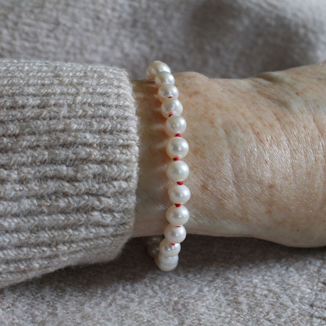 A close-up of a person's wrist wearing a  white pearl bracelet hand -knotted on ruby silk, paired with a beige ribbed knit sweater.