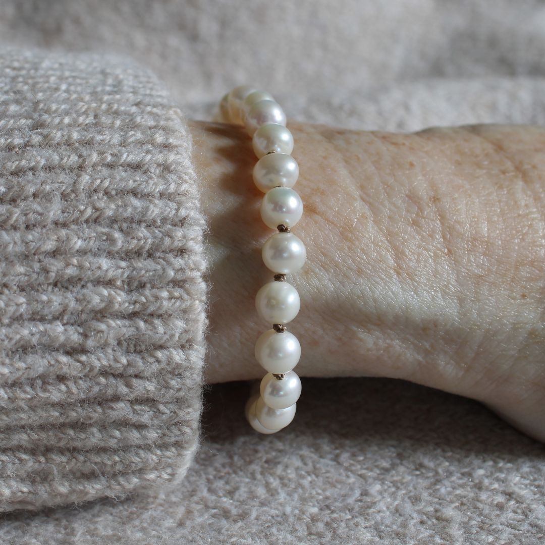 A close-up of a person's wrist wearing a bracelet made of white freshwater pearls, with a beige knit sweater cuff visible on the left.