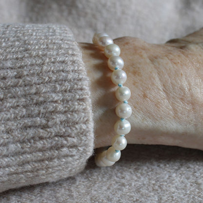 A close-up of a person's wrist wearing a pearl bracelet hand-knotted with turquoise silk, paired with a beige knit sweater cuff.