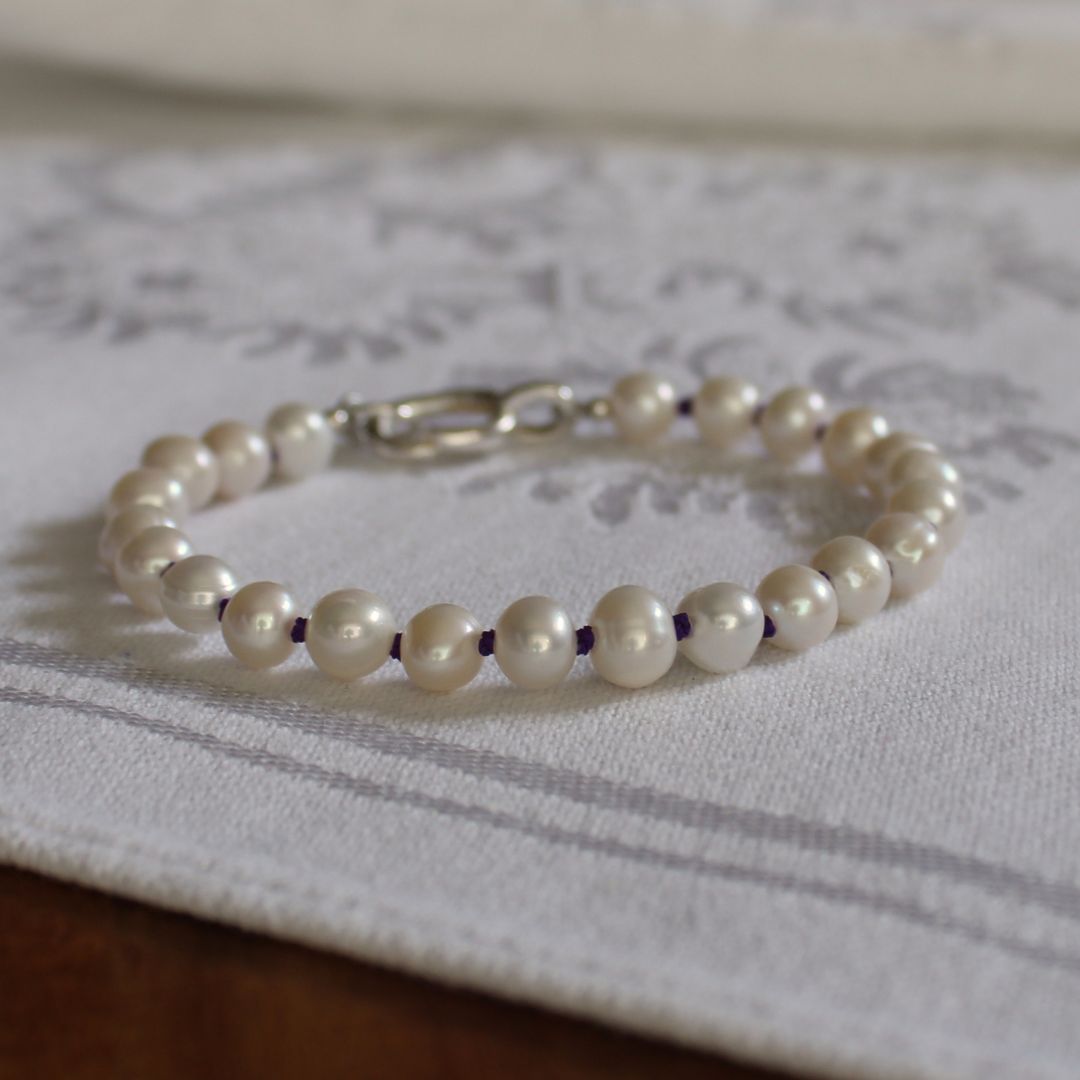 A beaded bracelet made of white pearls, hand-knotted on amethyst silk, with a silver oval charm clasp, resting on a white patterned fabric surface.