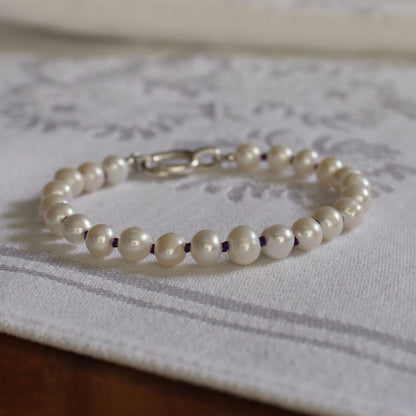 A beaded bracelet made of white pearls, hand-knotted on amethyst silk, with a silver oval charm clasp, resting on a white patterned fabric surface.