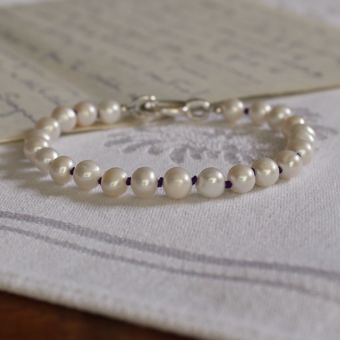 A bracelet made of white pearls hand-knotted on amethyst silk, with a silver clasp, resting on a white embroidered fabric surface.