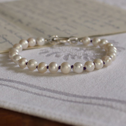 A bracelet made of white pearls hand-knotted on amethyst silk, with a silver clasp, resting on a white embroidered fabric surface.