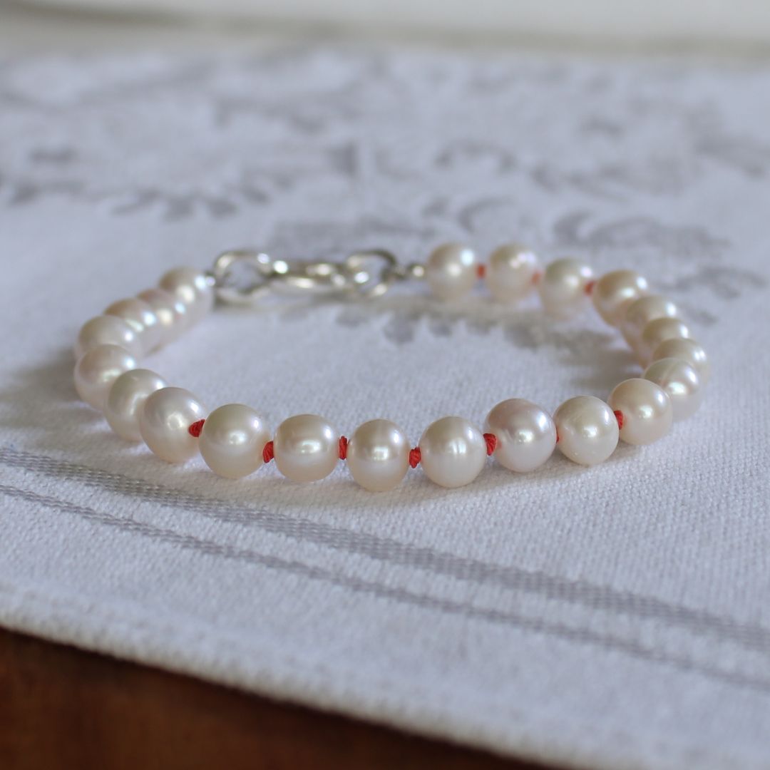A bracelet made of white freshwater pearls hand-knotted with coral silk, with a silver oval charmer clasp, resting on a white patterned fabric surface.