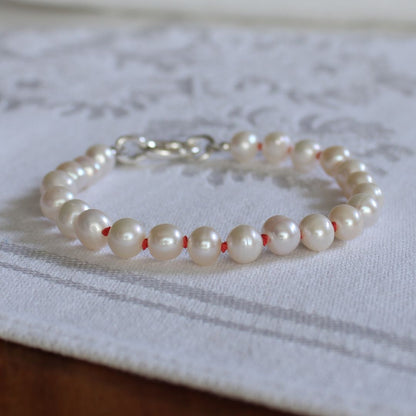 A bracelet made of white freshwater pearls hand-knotted with coral silk, with a silver oval charmer clasp, resting on a white patterned fabric surface.