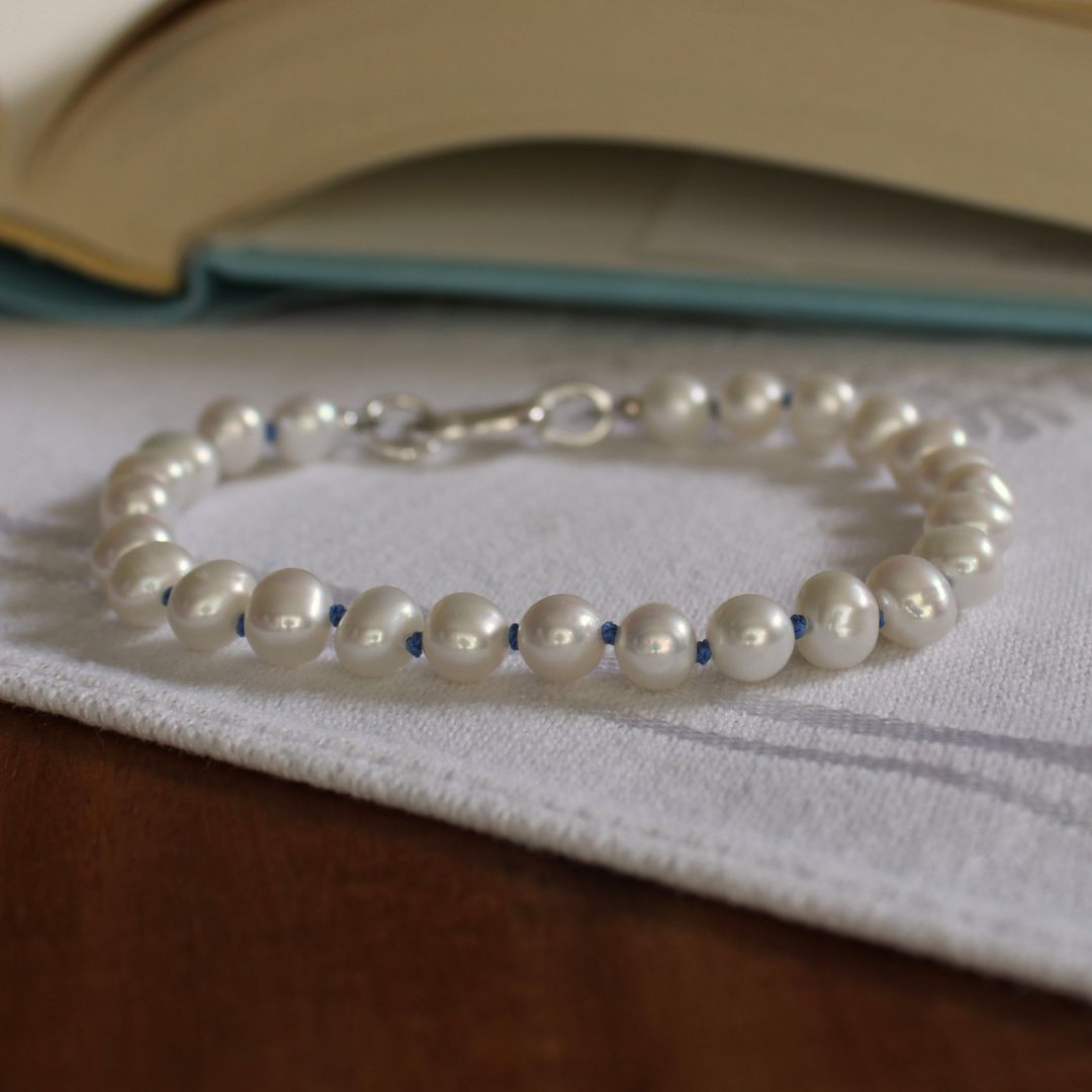 A beaded bracelet made of white pearls knotted on delft blue silk and a silver clasp, resting on a white textured cloth over a wooden surface with an open book blurred in the background.