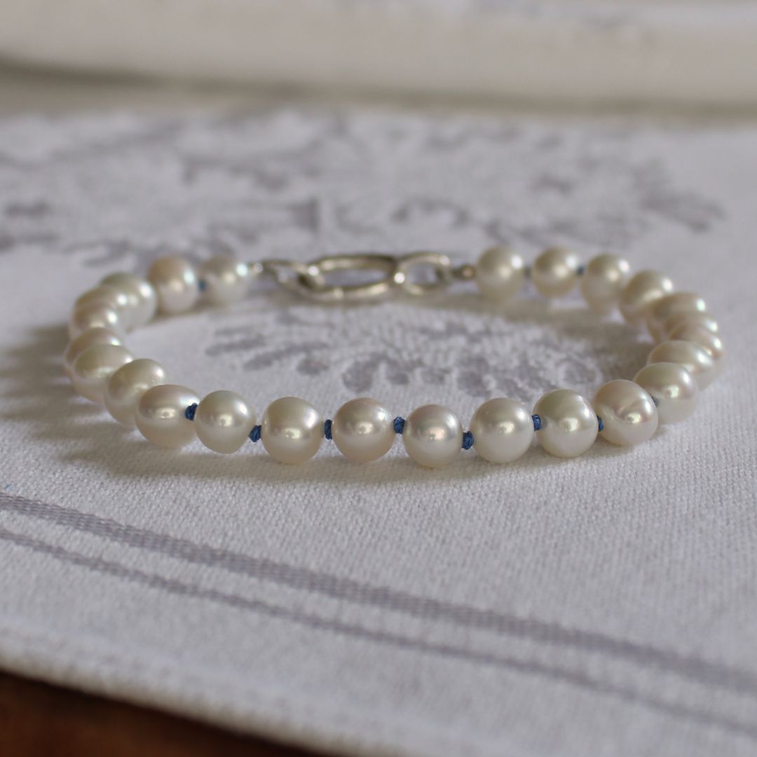 A beaded bracelet made of white pearls hand-knotted with delft blue silk, with a silver clasp, resting on a white patterned fabric surface.
