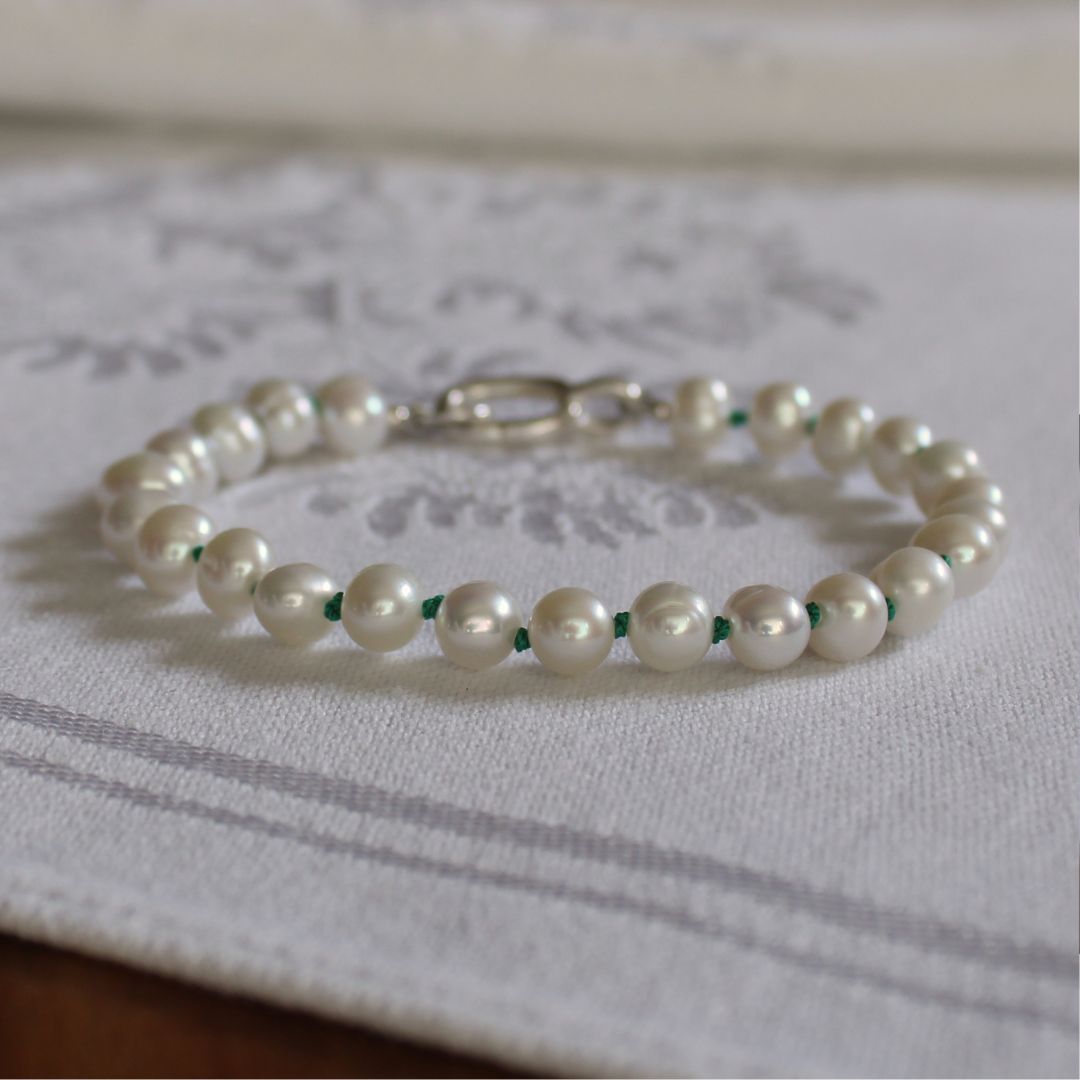 A beaded bracelet made of white freshwater pearls knotted on emerald green silk, with a silver clasp, resting on a white patterned fabric surface.