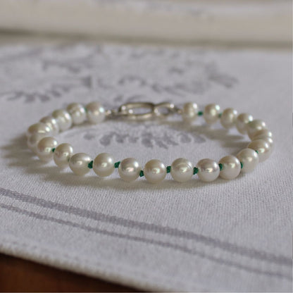A beaded bracelet made of white freshwater pearls knotted on emerald green silk, with a silver clasp, resting on a white patterned fabric surface.