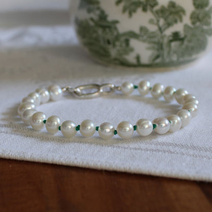 A beaded bracelet made of white pearls knotted on emerald green silk, with a silver clasp, resting on a white textured cloth over a wooden surface.