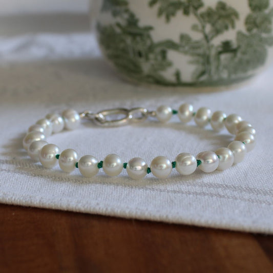 A beaded bracelet made of white pearls knotted on emerald green silk, with a silver clasp, resting on a white textured cloth over a wooden surface.