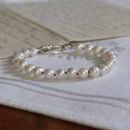 A pearl beaded bracelet hand-knotted with garnet silk, and with a silver oval charmer clasp, resting on a white embroidered cloth with vintage handwritten letters in the background.