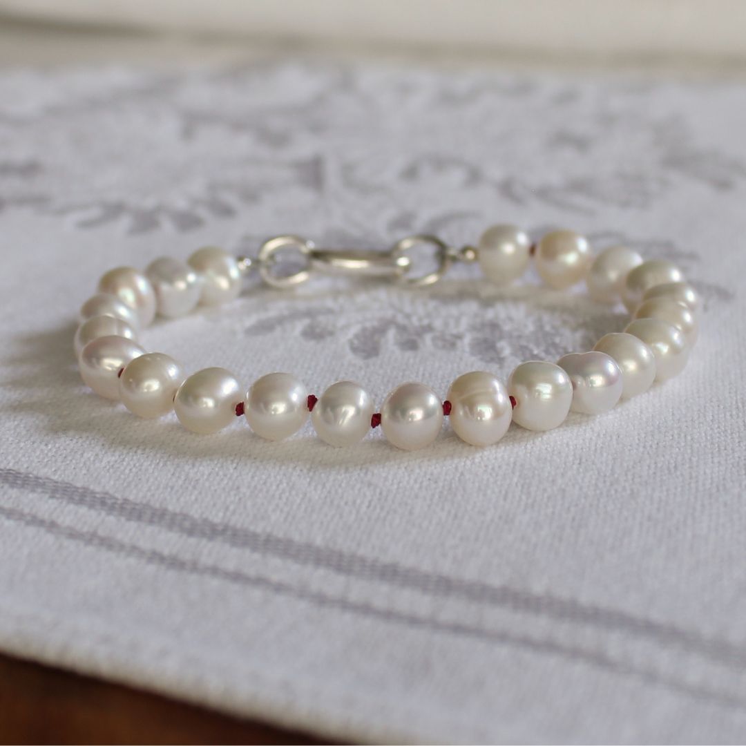 A freshwater pearl bracelet, hand-knotted with garnet silk, with a silver oval charmer clasp, resting on a white patterned fabric surface.