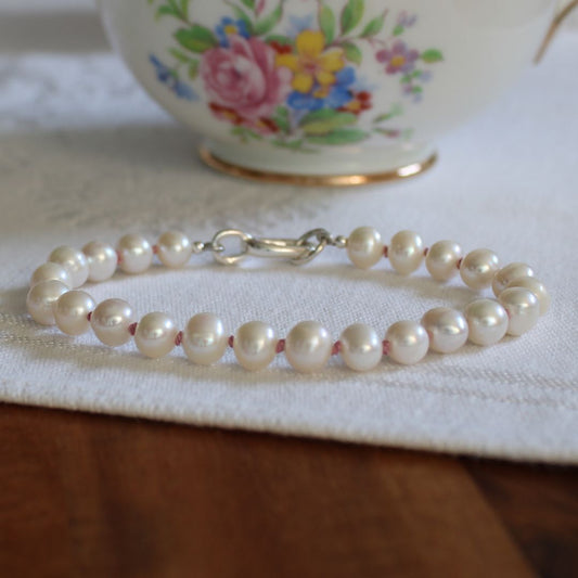 A strand bracelet made of white freshwater pearls, knotted with old rose silk, with a silver clasp, resting on a white textured fabric runner with a wooden table surface below and a floral teacup in the blurred background.