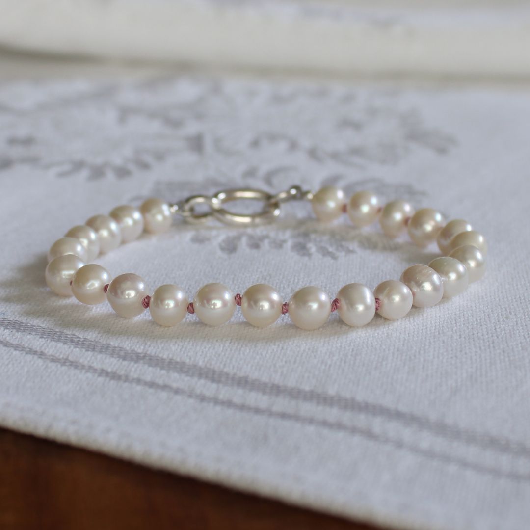 A strand bracelet made of white freshwater pearls with a silver clasp, resting on a white textured fabric runner.