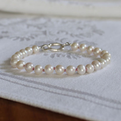 A strand bracelet made of white freshwater pearls with a silver clasp, resting on a white textured fabric runner.