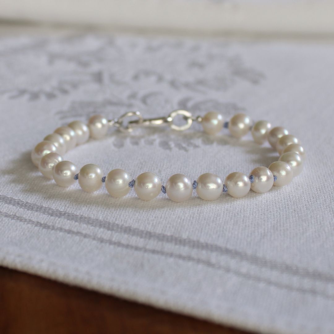 A bracelet made of white pearls, knotted on periwinkle silk, with a silver clasp, resting on a white embroidered fabric surface.