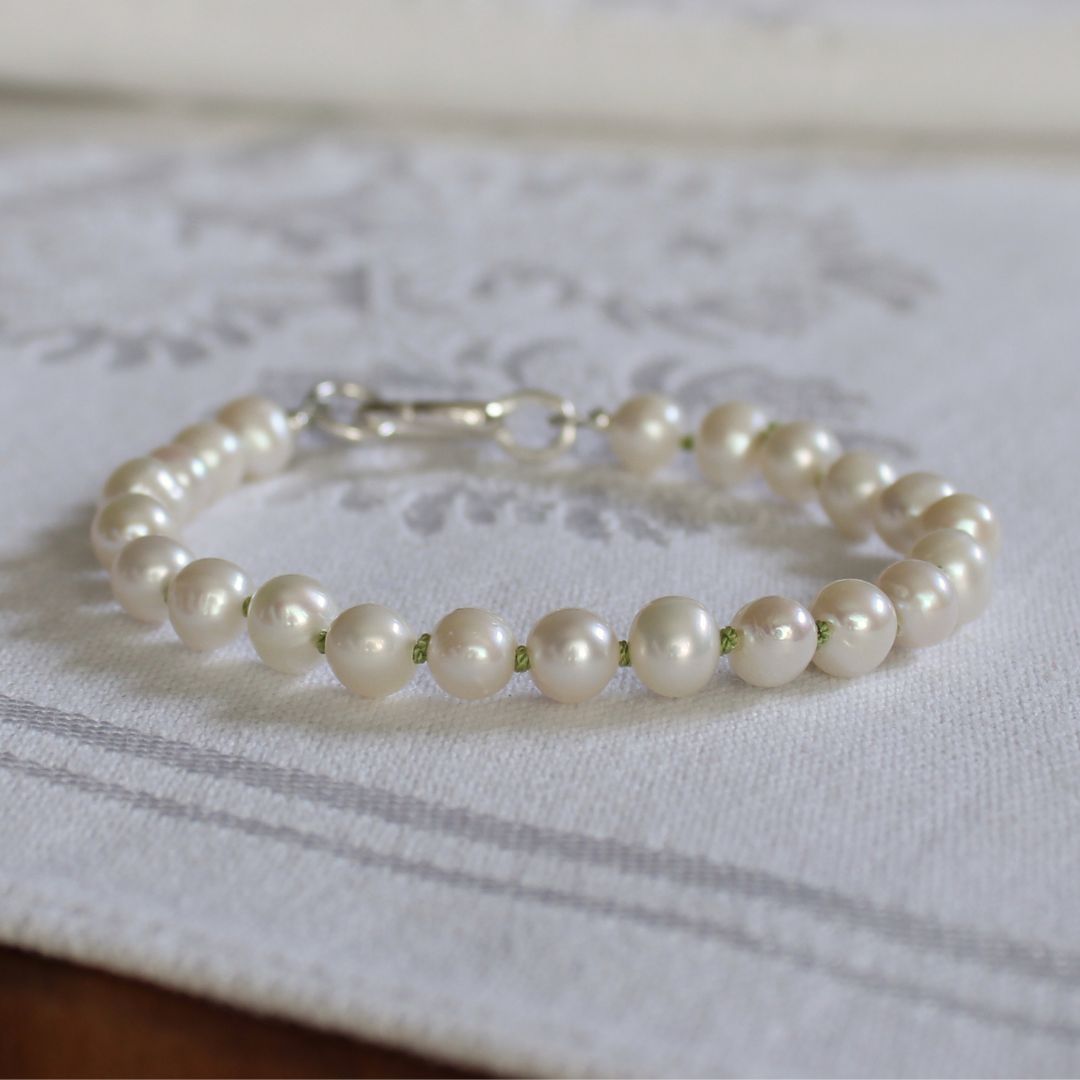 A bracelet made of white freshwater pearls knotted in peridot silk, finished with a silver oval charmer clasp, resting on a white embroidered fabric surface.