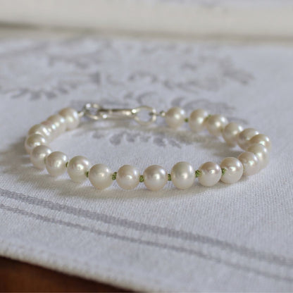 A bracelet made of white freshwater pearls knotted in peridot silk, finished with a silver oval charmer clasp, resting on a white embroidered fabric surface.