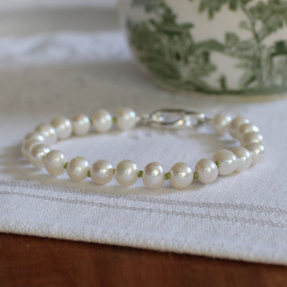 A bracelet made of white freshwater pearls hand-knotted in peridot silk, featuring a silver oval charmer clasp, resting on a white textured cloth.