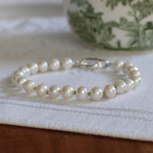 A bracelet made of white freshwater pearls hand-knotted in peridot silk, featuring a silver oval charmer clasp, resting on a white textured cloth.