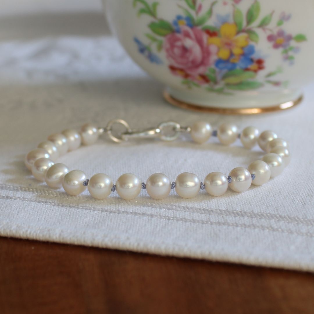 A white pearl bracelet, hand-knotted with periwinkle silk, with a silver oval charmer clasp, resting on a white textured fabric surface with a floral ceramic cup blurred in the background.