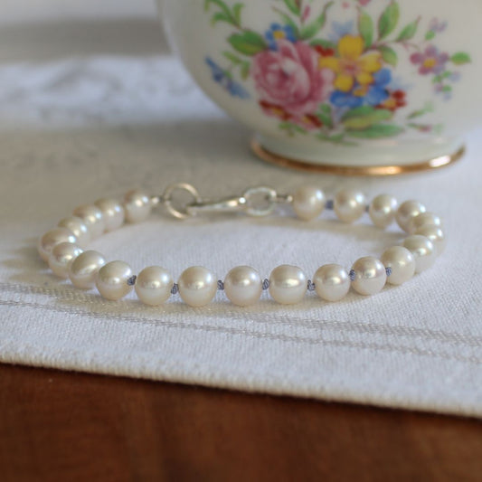 A white pearl bracelet, hand-knotted with periwinkle silk, with a silver oval charmer clasp, resting on a white textured fabric surface with a floral ceramic cup blurred in the background.