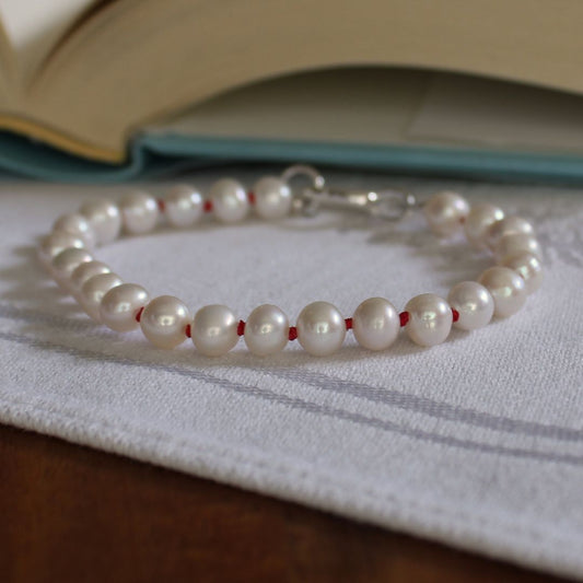 A pearl bracelet hand-knotted with ruby silk, with a silver clasp, resting on a white textured cloth with an open book in the background.
