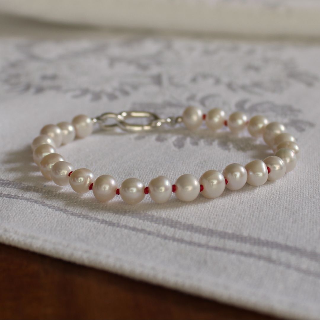 A beaded bracelet made of glossy white freshwater pearls hand-knotted on ruby silk, with a silver charmer clasp, resting on a white patterned fabric surface.