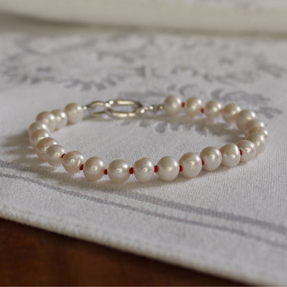 A beaded bracelet made of glossy white freshwater pearls hand-knotted on ruby silk, with a silver charmer clasp, resting on a white patterned fabric surface.