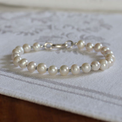 A pearl bracelet knotted with silver silk, with a silver clasp resting on a white patterned fabric surface.
