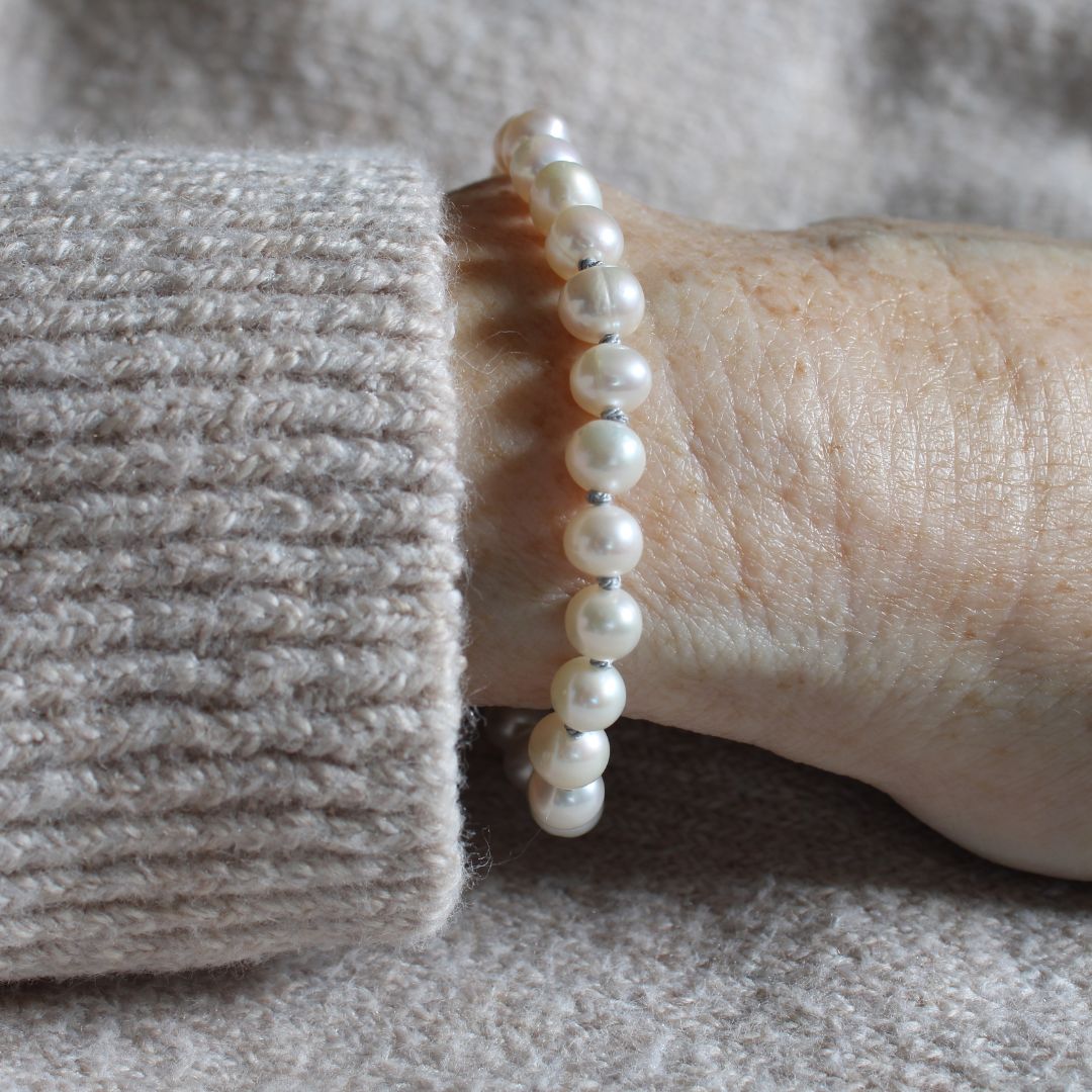 A close-up of a person's wrist wearing a bracelet of  freshwater pearls hand-knotted with silver silk, shown with a beige knit sweater cuff against a soft beige fabric background.